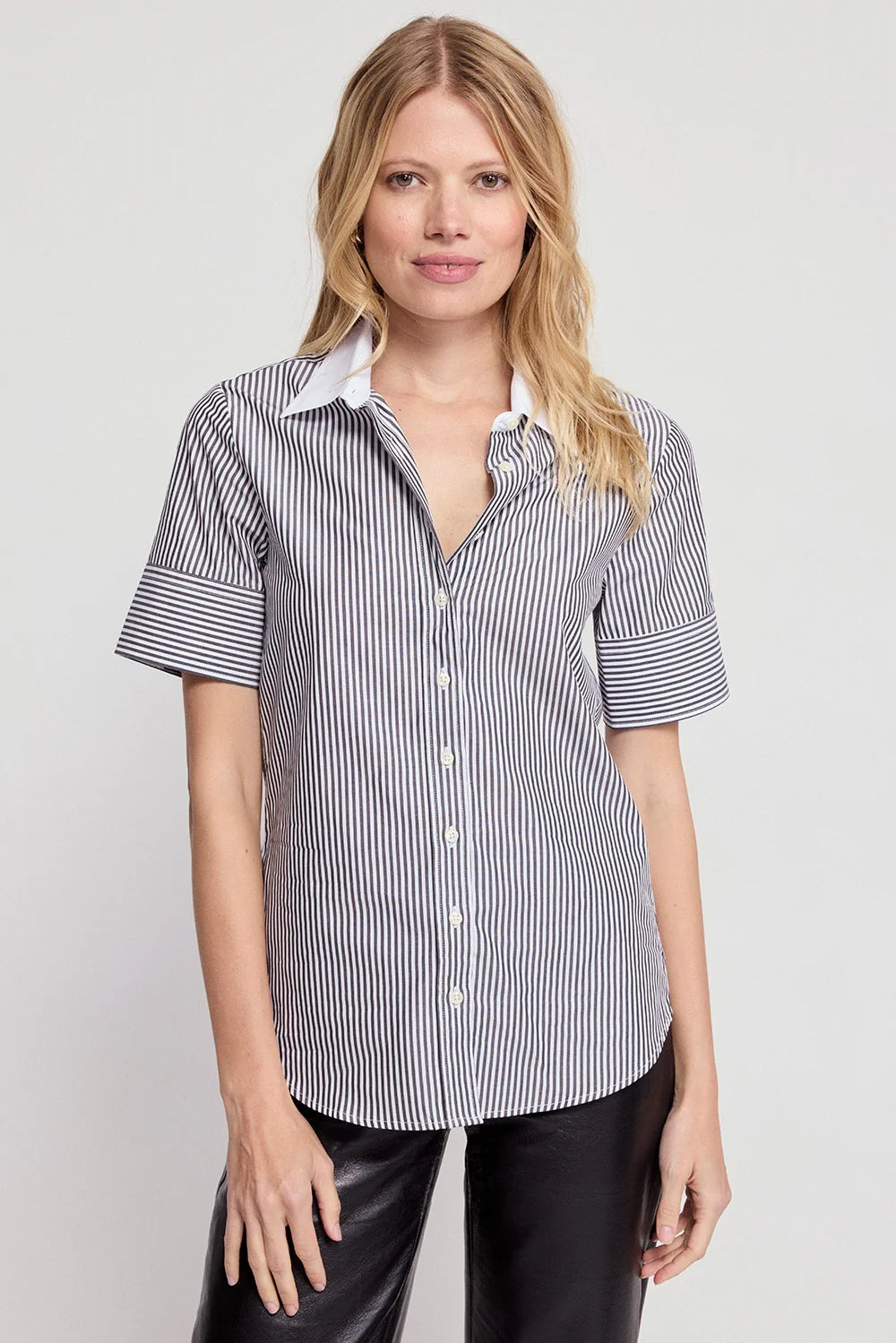 A woman with blonde hair wears THE SHIRT's Short Sleeve Shirt in cotton stretch with black-and-white vertical stripes and No Gape button technology, paired with black pants, against a plain light background, smiling slightly.