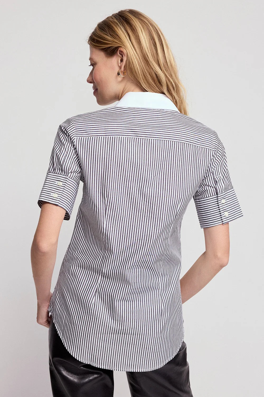 A woman with long blonde hair is seen from the back, wearing THE SHIRT's The Short Sleeve Shirt in black and white vertical stripes with black pants, against a light grey background.