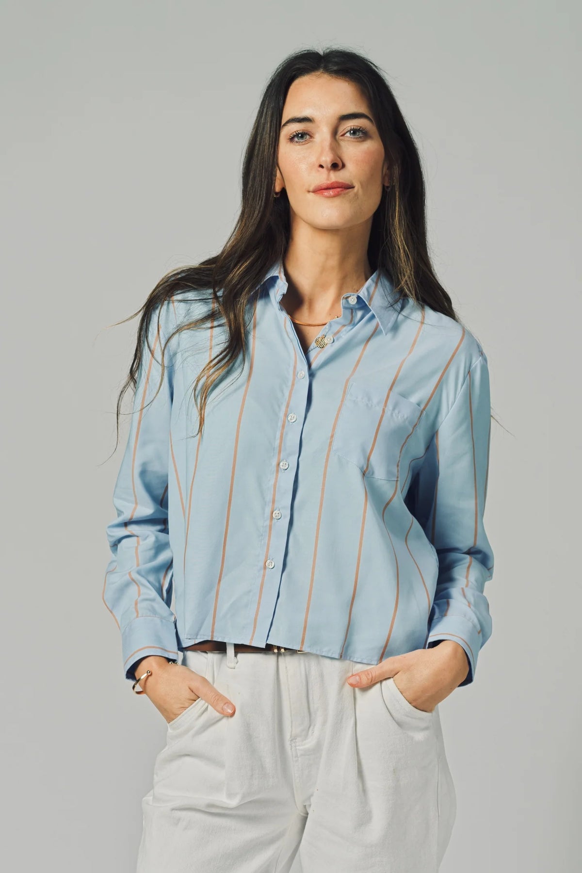 A woman with long dark hair wears THE SHIRT’s The LS Boxy Crop in light blue stripes and white pants, standing with her hands in her pockets against a plain gray background.