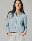 A woman with long dark hair wears THE SHIRT’s The LS Boxy Crop in light blue stripes and white pants, standing with her hands in her pockets against a plain gray background.
