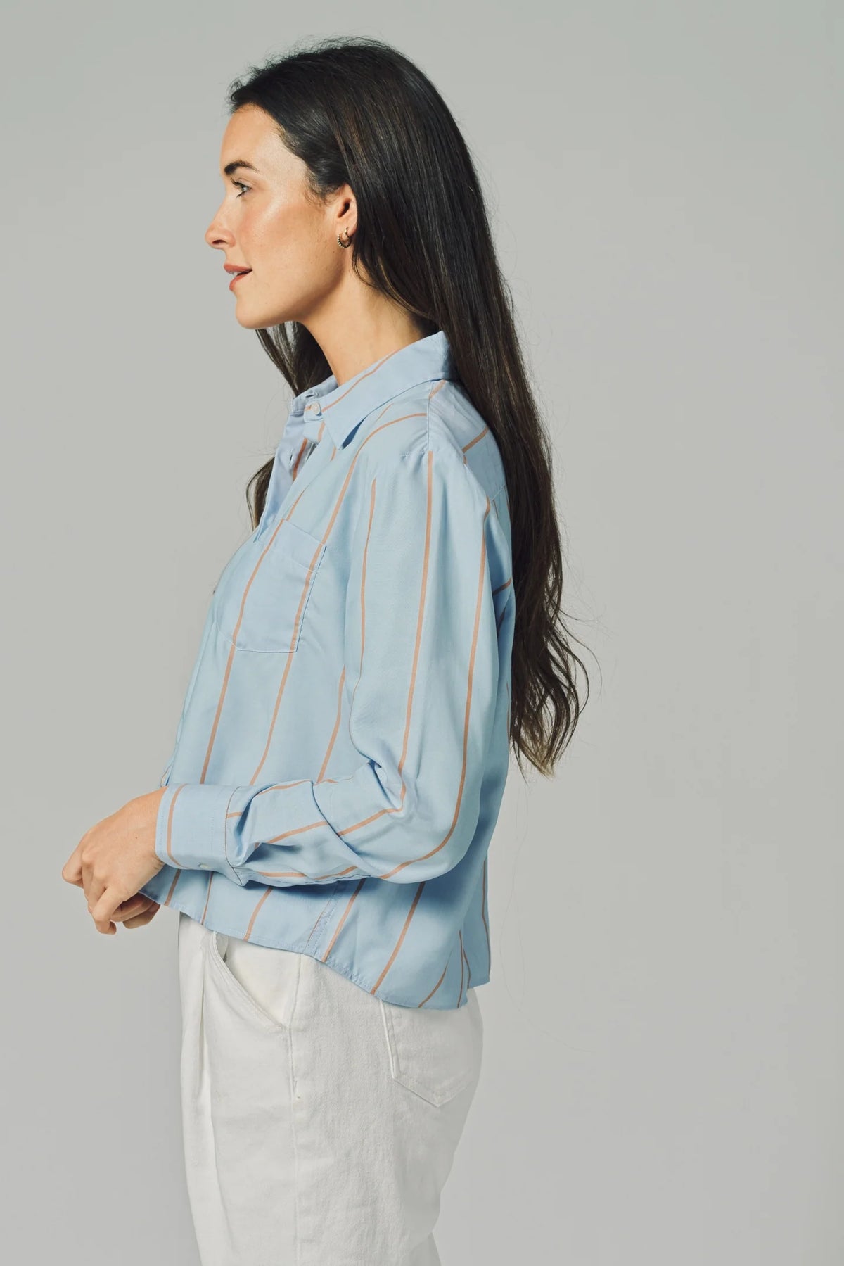 A woman with long dark hair, wearing THE SHIRT's The LS Boxy Crop— a light blue, thin-striped button-up with No Gape technology—and white pants, stands in profile against a plain gray background.