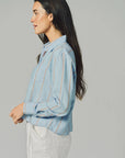 A woman with long dark hair, wearing THE SHIRT's The LS Boxy Crop— a light blue, thin-striped button-up with No Gape technology—and white pants, stands in profile against a plain gray background.
