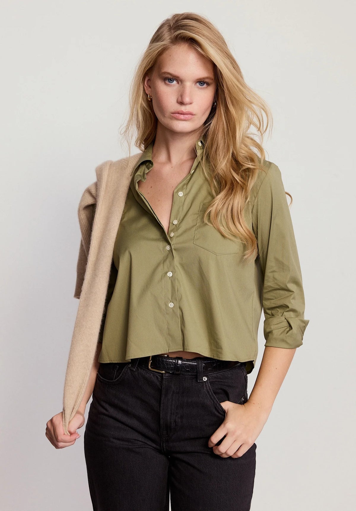 A woman with long blonde hair wears THE SHIRT’s The LS Boxy Crop in olive green, paired with black jeans and a beige sweater draped over her shoulder, standing against a plain light background.