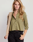 A woman with long blonde hair wears THE SHIRT’s The LS Boxy Crop in olive green, paired with black jeans and a beige sweater draped over her shoulder, standing against a plain light background.