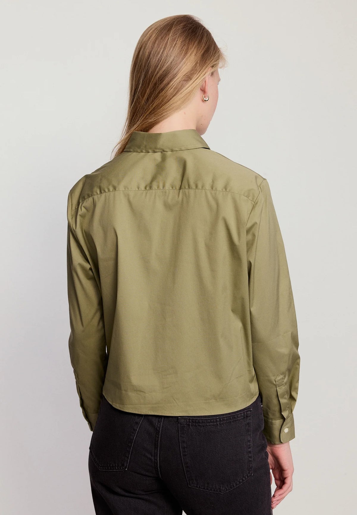 A woman with long blonde hair, seen from behind, wears THE SHIRT’s The LS Boxy Crop in olive green with the No Gape button and dark high-waisted jeans against a plain light background.