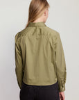A woman with long blonde hair, seen from behind, wears THE SHIRT’s The LS Boxy Crop in olive green with the No Gape button and dark high-waisted jeans against a plain light background.