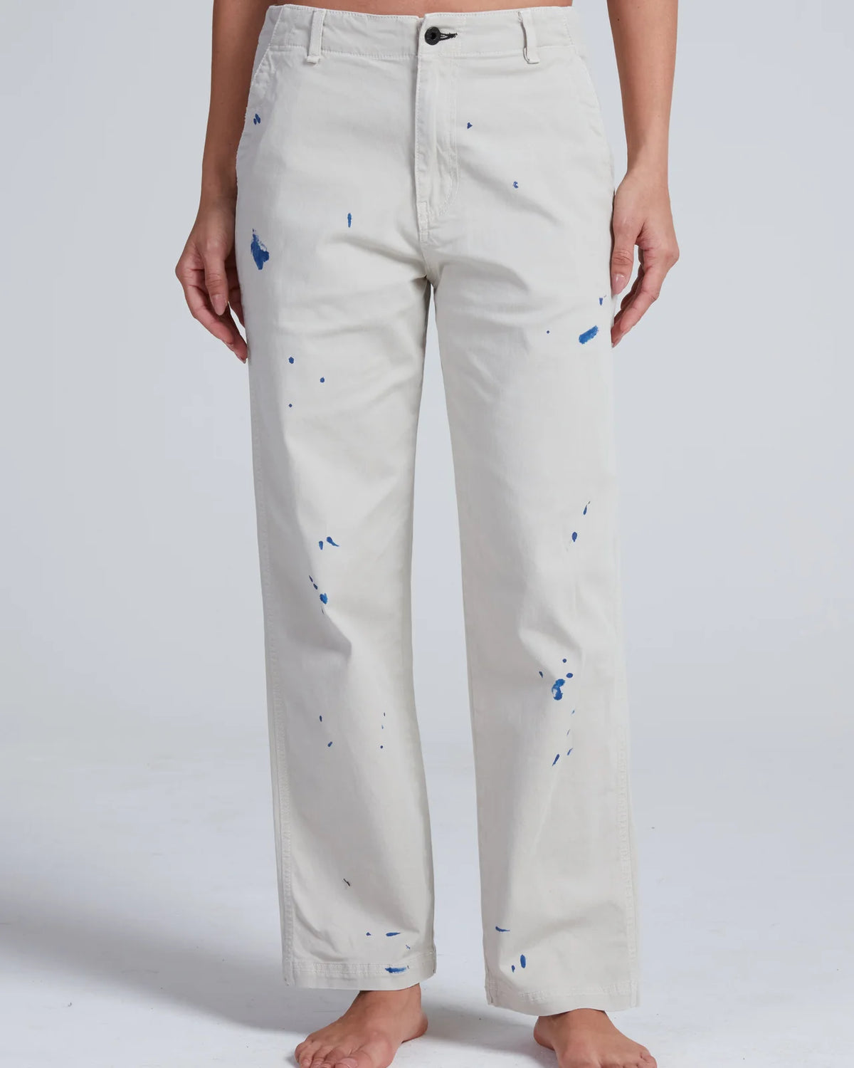 A person stands barefoot, shown from the waist down against a plain light background, wearing ASKK’s Tomboy Twill high-waist straight-leg pants in light beige with blue paint splatters, crafted from stretch twill.
