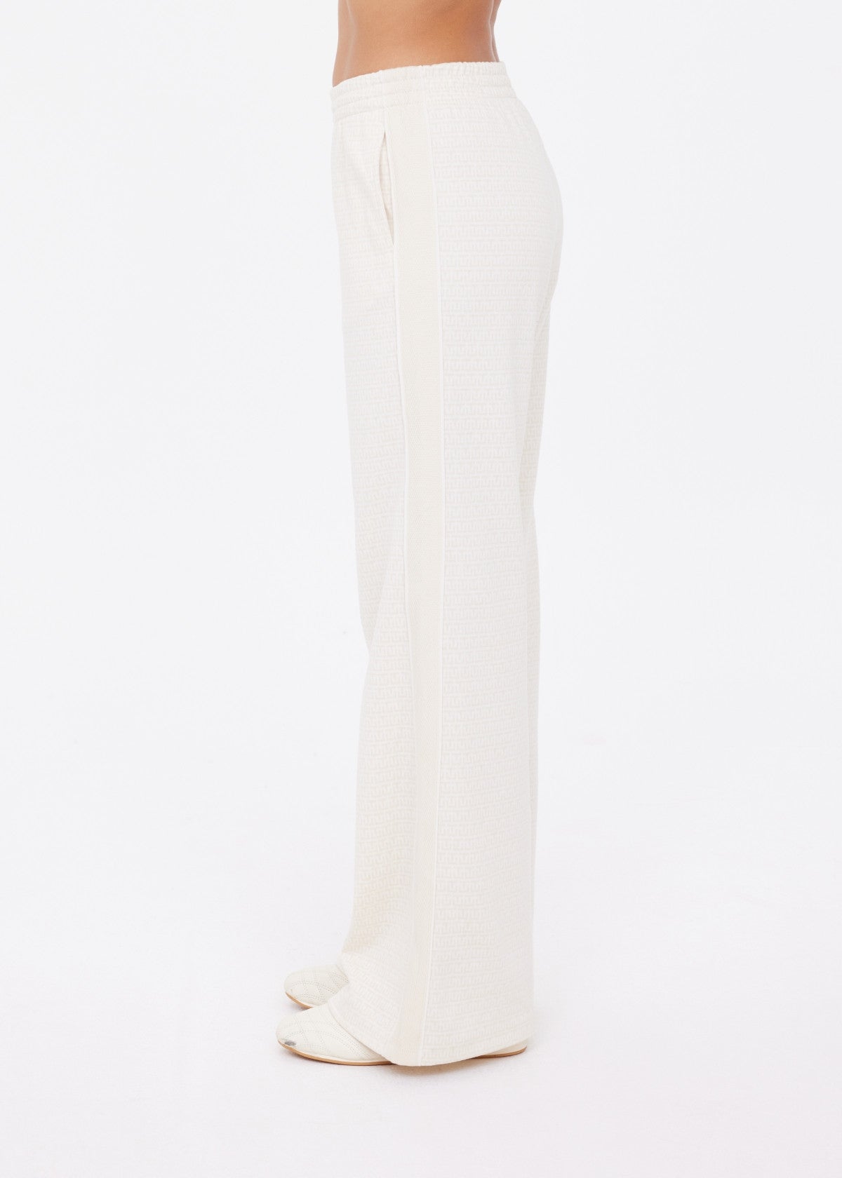 A person is shown standing sideways against a plain white background, wearing The Upside’s NAVAR MARNIE WIDE LEG PANT in cream paired with open-toe sandals.