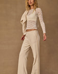 A woman with long wavy blonde hair poses against a tan background, wearing The Upside's NAVAR MARNIE WIDE LEG PANT, a sheer white lace long-sleeve top over a beige camisole, and has a beige sweater draped over her shoulders.