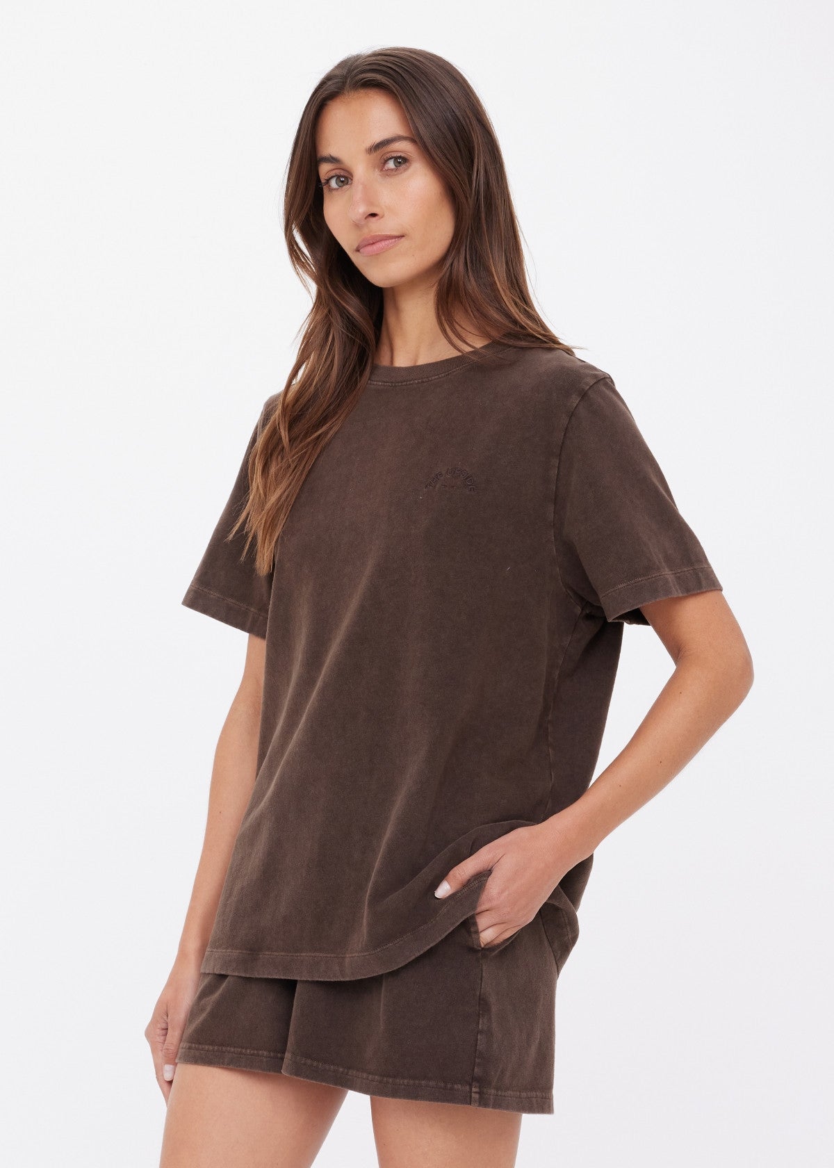 A woman wears the Citizens Of Humanity/AGOLDE SOHO EVA CREW TEE in deep cocoa brown with matching brushed recycled cotton shorts, standing against a white background with one hand in her pocket and a neutral expression.