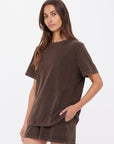 A woman wears the Citizens Of Humanity/AGOLDE SOHO EVA CREW TEE in deep cocoa brown with matching brushed recycled cotton shorts, standing against a white background with one hand in her pocket and a neutral expression.