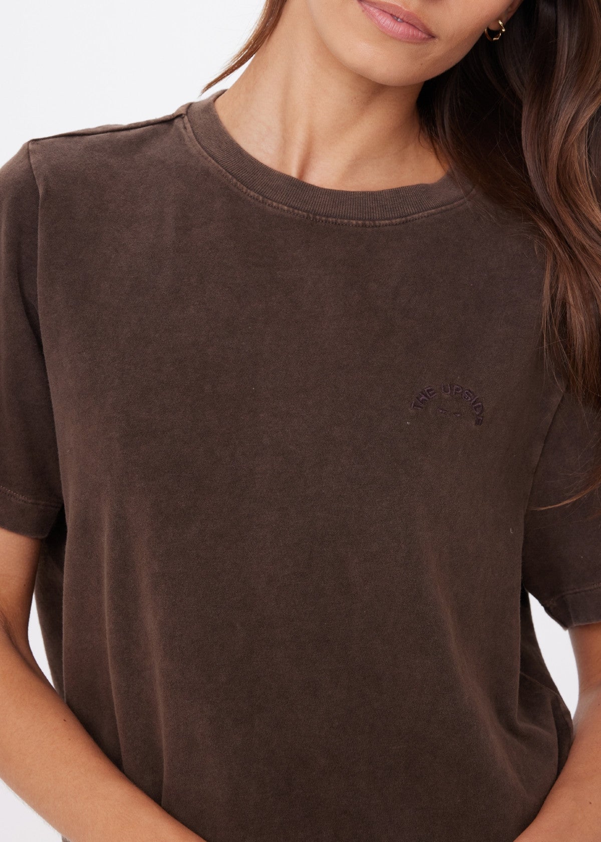 A woman with long, wavy brown hair wears the SOHO EVA CREW TEE by Citizens Of Humanity/AGOLDE—a loose, deep cocoa brown crew neck made from brushed recycled cotton with subtle embroidery on the left chest. She is partially cropped out of frame.