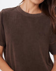 A woman with long, wavy brown hair wears the SOHO EVA CREW TEE by Citizens Of Humanity/AGOLDE—a loose, deep cocoa brown crew neck made from brushed recycled cotton with subtle embroidery on the left chest. She is partially cropped out of frame.