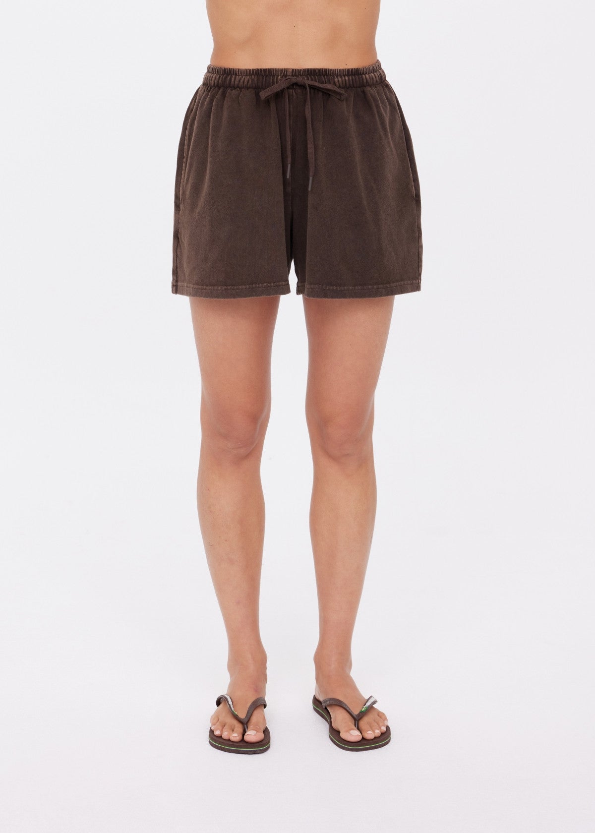 A person wears The Upside's SOHO DOLCI SHORT in dark brown, crafted from recycled cotton, with black flip-flops. Only their legs and lower torso are visible against a plain white background.