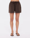 A person wears The Upside's SOHO DOLCI SHORT in dark brown, crafted from recycled cotton, with black flip-flops. Only their legs and lower torso are visible against a plain white background.