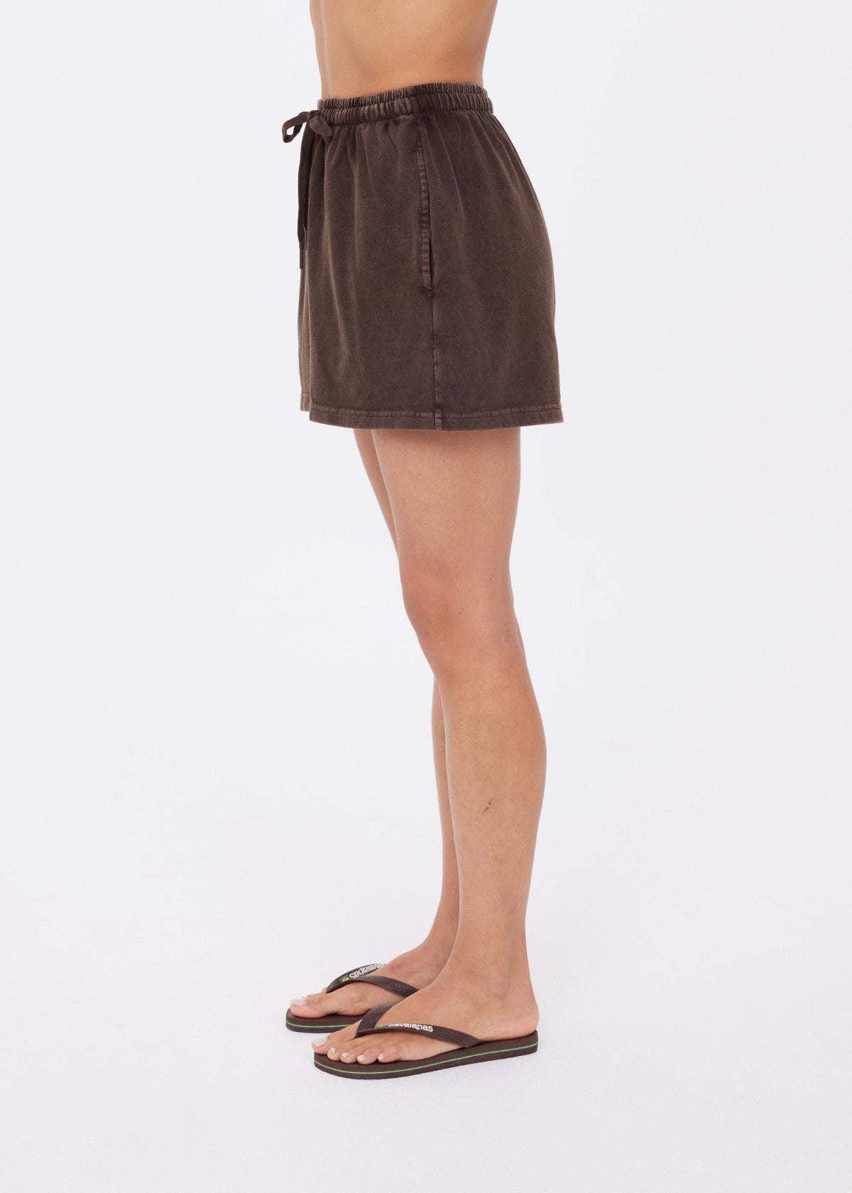 A person in profile wears The Upside SOHO DOLCI SHORT mini skirt in brown, crafted from recycled cotton, and black flip-flops, set against a plain white background.