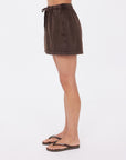 A person in profile wears The Upside SOHO DOLCI SHORT mini skirt in brown, crafted from recycled cotton, and black flip-flops, set against a plain white background.