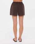 A person stands facing away in The Upside's SOHO DOLCI SHORT, made from brown recycled cotton, paired with black sandals against a plain white background.