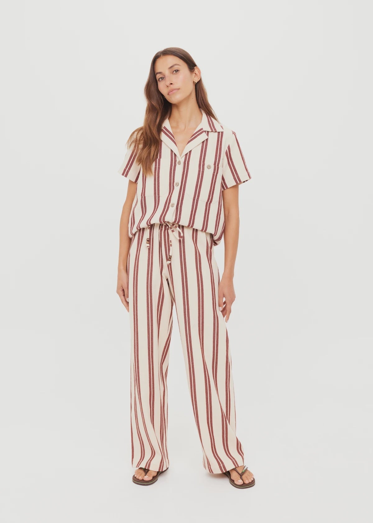 A woman stands against a plain white background wearing The Upside's cream short-sleeve button-up shirt and BAJA CHARLI DRAWSTRING PANT in burnt red stripes, paired with sandals.