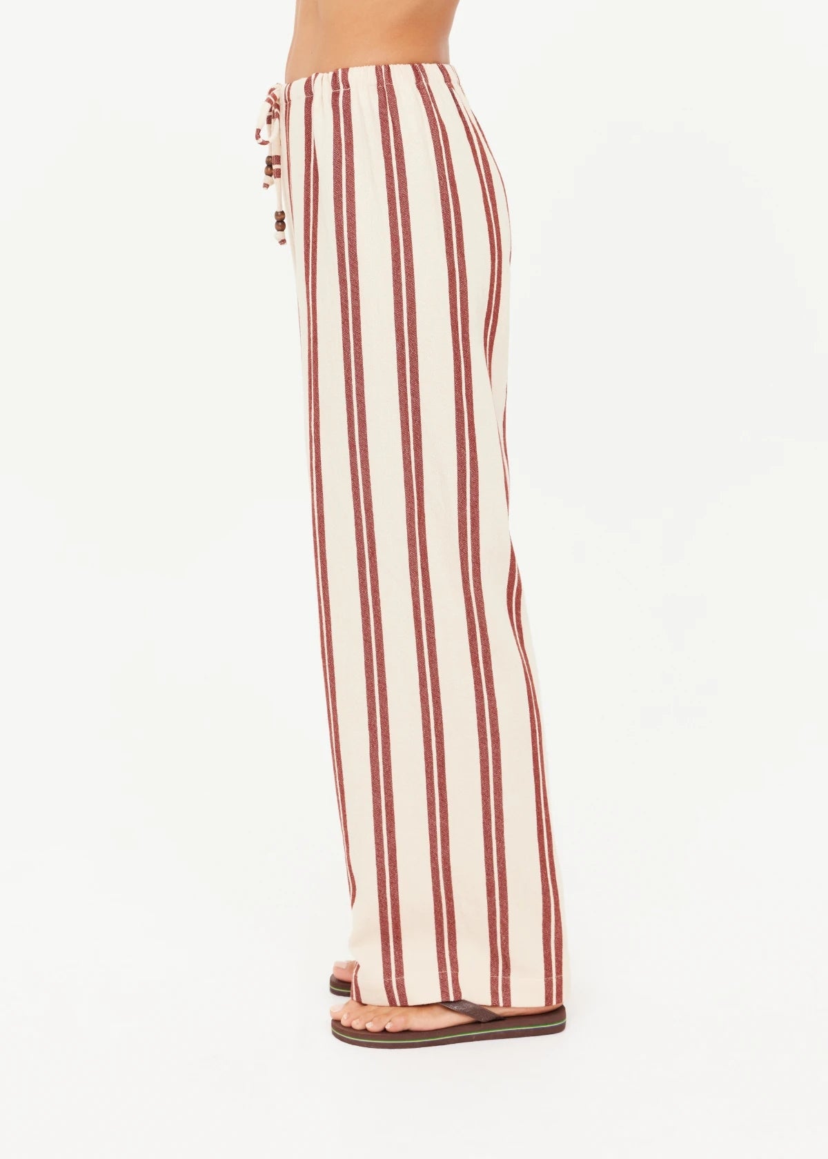 A person stands in profile against a plain white background, wearing The Upside's BAJA CHARLI DRAWSTRING PANT—high-waisted, straight leg pants with vertical burnt red stripes and a drawstring waist—paired with flat sandals.