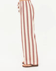 A person stands in profile against a plain white background, wearing The Upside's BAJA CHARLI DRAWSTRING PANT—high-waisted, straight leg pants with vertical burnt red stripes and a drawstring waist—paired with flat sandals.