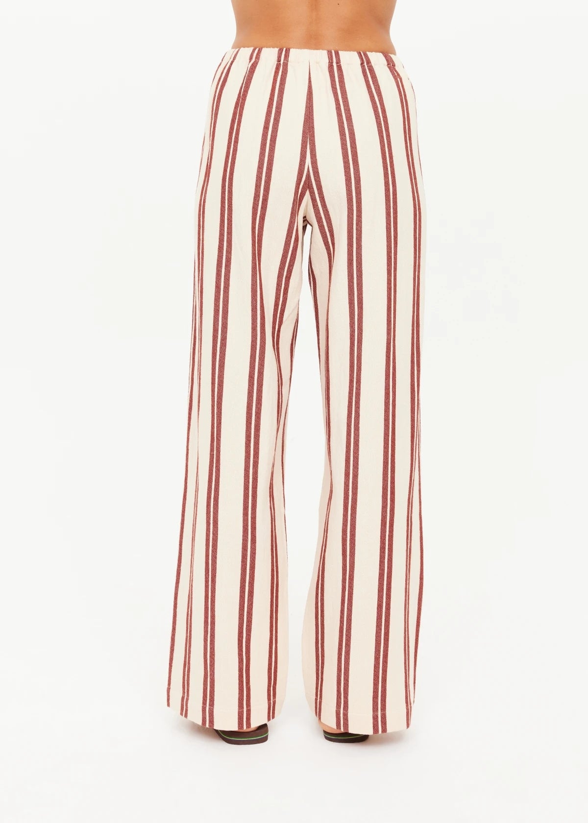 A person is shown from the back wearing The Upside's BAJA CHARLI DRAWSTRING PANT, featuring high-waisted, straight legs with vertical burnt red and white stripes, set against a plain white background.