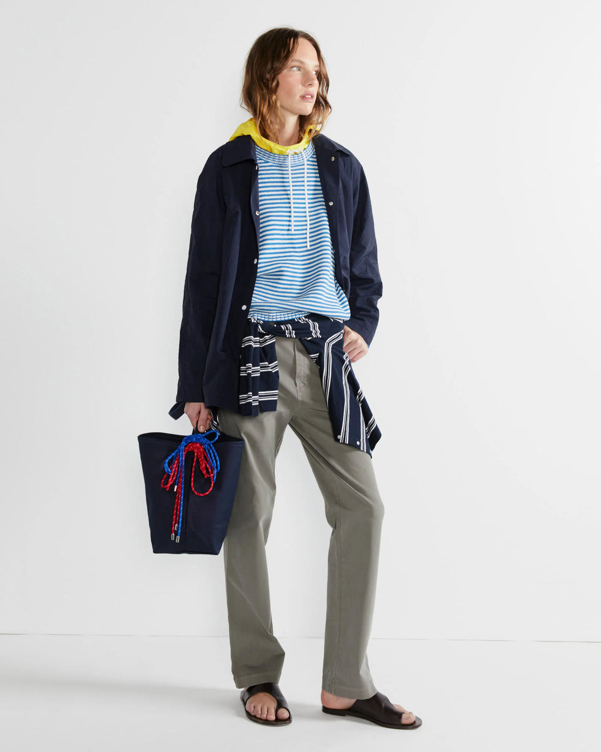 Wearing Kule's "The Uma" yellow hooded top under a blue striped shirt and navy jacket, with khaki pants, black sandals, a navy cotton canvas tote with rope handles, and a plaid shirt tied at the waist, the person stands against a plain background.