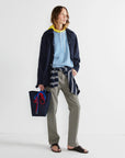Wearing Kule's "The Uma" yellow hooded top under a blue striped shirt and navy jacket, with khaki pants, black sandals, a navy cotton canvas tote with rope handles, and a plaid shirt tied at the waist, the person stands against a plain background.