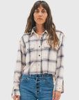 A woman with long brown hair wears the ASKK Crosby Flannel, a white and blue plaid button-up shirt, paired with high-waisted blue jeans, standing facing forward against a plain light background.