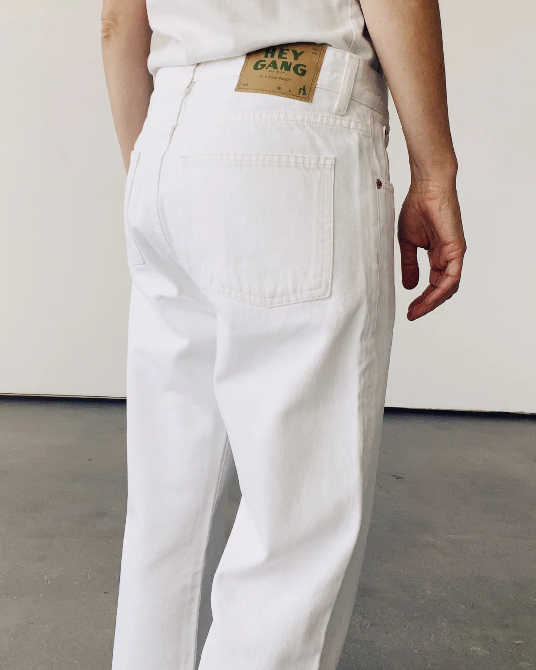A person wears Hey Gang's The Loose Crop white mid-rise jeans, paired with a matching shirt, facing away from the camera. The 100% cotton denim jeans feature a green and brown label on the waistband. They stand against a plain light-colored wall and floor.