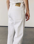 A person wears Hey Gang's The Loose Crop white mid-rise jeans, paired with a matching shirt, facing away from the camera. The 100% cotton denim jeans feature a green and brown label on the waistband. They stand against a plain light-colored wall and floor.