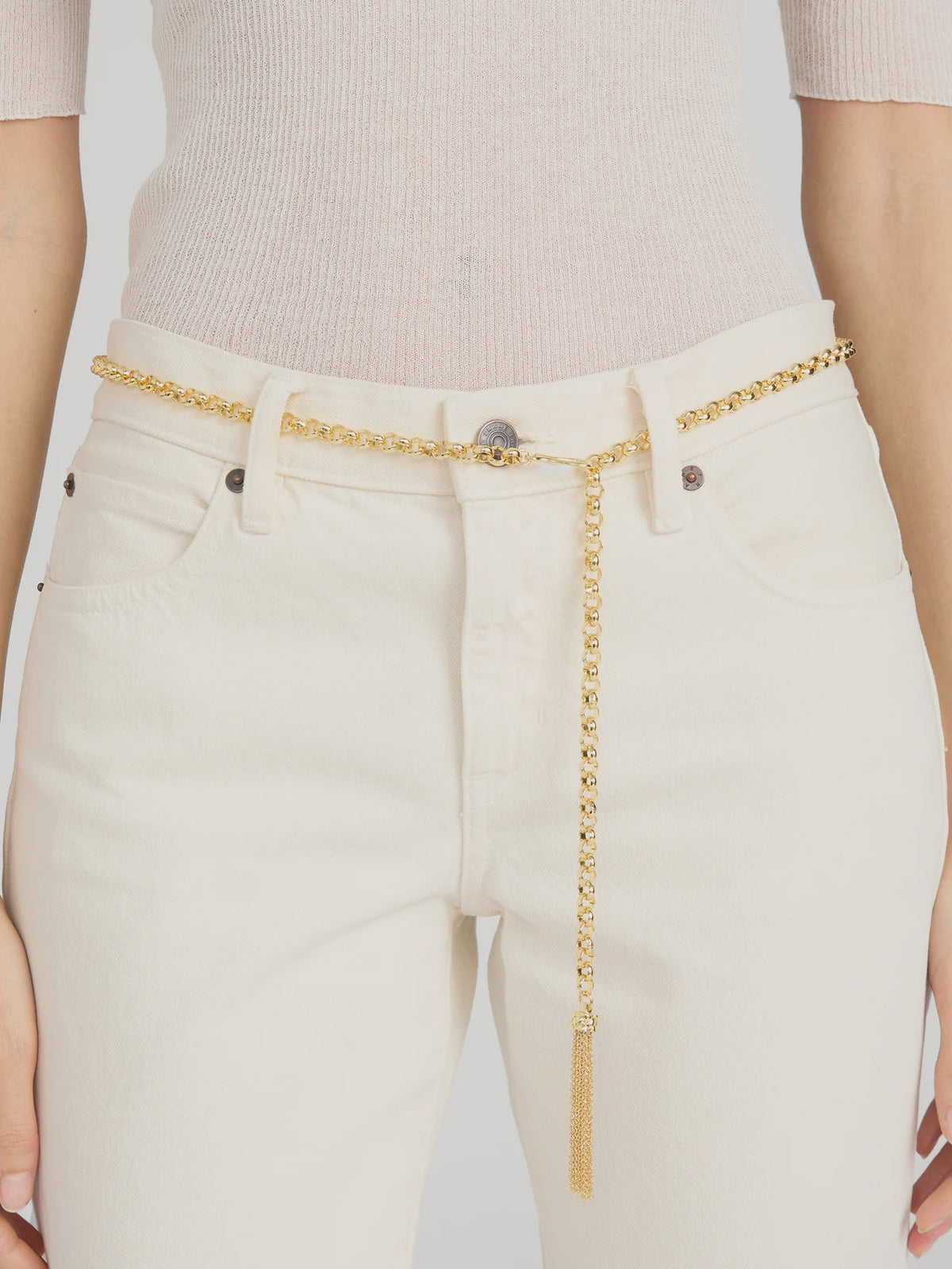 A person wears high-waisted, off-white pants with The Sebastian Belt Gold by FRAME looped through the belt loops and hanging down one side with a tassel detail, paired with a light beige ribbed top.