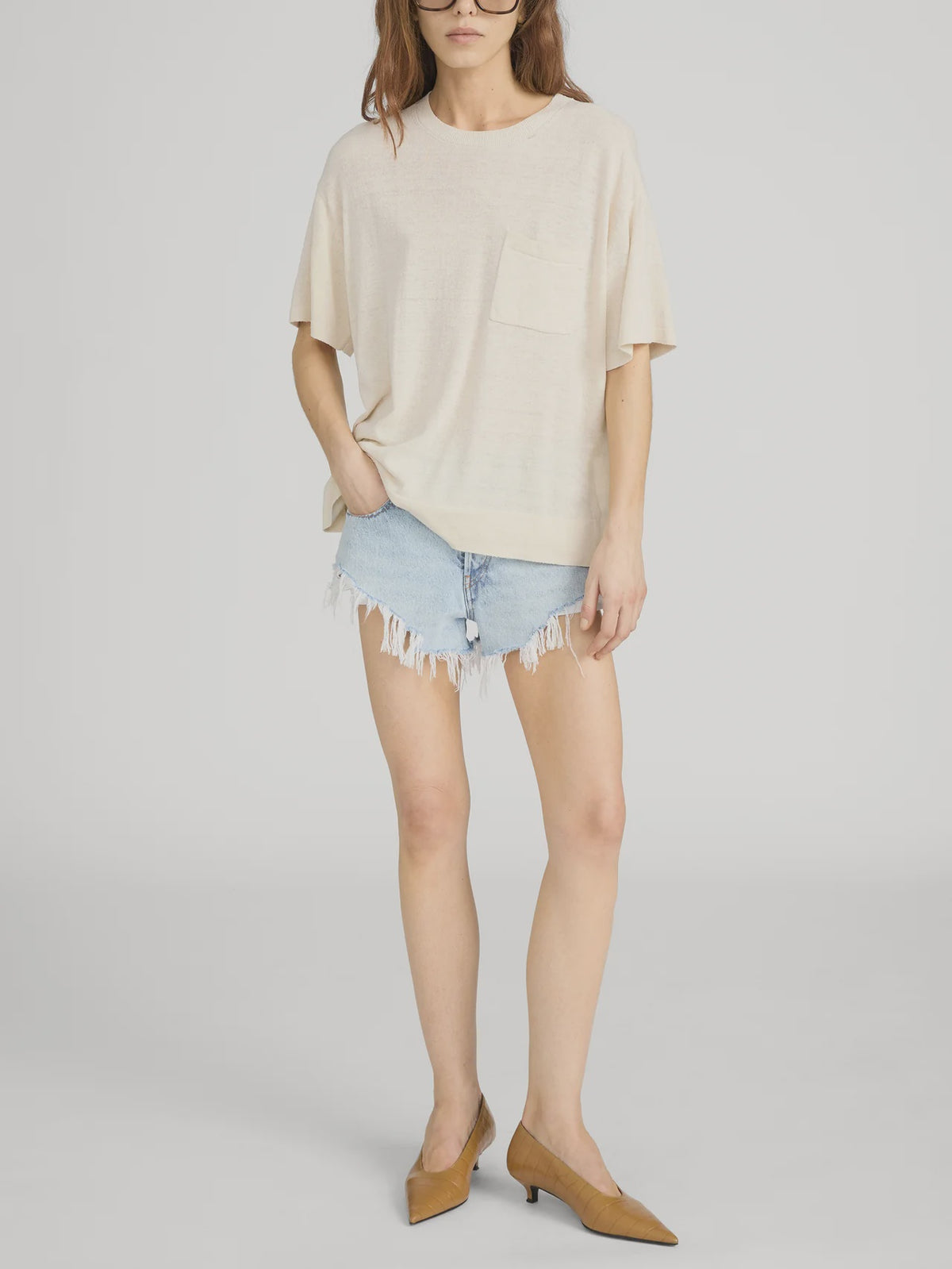 Someone stands against a plain light background wearing FRAME's The Knit Plus One Tee, a relaxed fit beige crewneck with a front pocket, paired with light blue frayed denim shorts and brown low-heeled shoes.