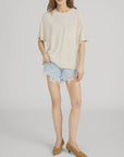 Someone stands against a plain light background wearing FRAME's The Knit Plus One Tee, a relaxed fit beige crewneck with a front pocket, paired with light blue frayed denim shorts and brown low-heeled shoes.