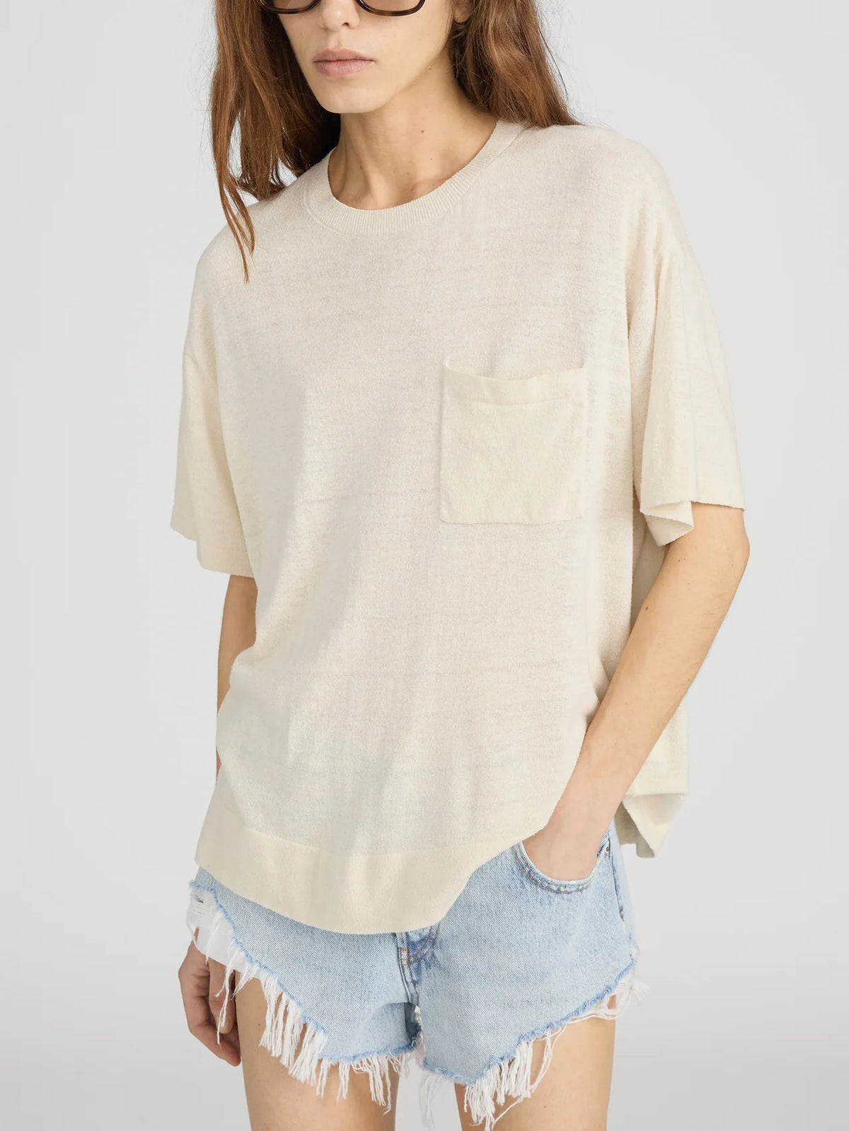 A woman wears the FRAME Knit Plus One Tee, a relaxed fit, short-sleeved beige crewneck with a chest pocket, paired with light blue frayed denim shorts. She stands against a plain background with one hand in her pocket.
