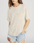 A woman wears the FRAME Knit Plus One Tee, a relaxed fit, short-sleeved beige crewneck with a chest pocket, paired with light blue frayed denim shorts. She stands against a plain background with one hand in her pocket.