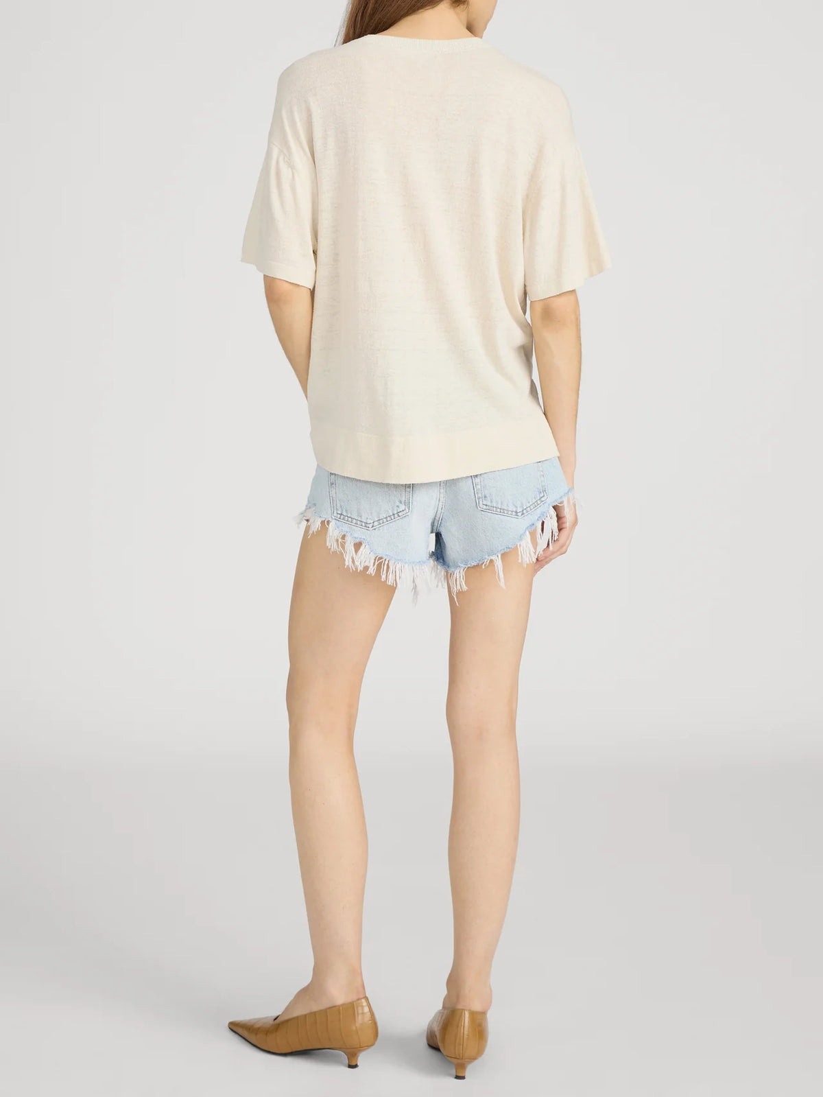 A person stands facing away in FRAME’s The Knit Plus One Tee— a relaxed fit, short-sleeve beige crewneck top—paired with light blue frayed denim shorts and tan low-heeled shoes against a plain light background.