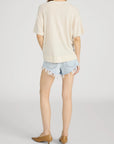 A person stands facing away in FRAME’s The Knit Plus One Tee— a relaxed fit, short-sleeve beige crewneck top—paired with light blue frayed denim shorts and tan low-heeled shoes against a plain light background.