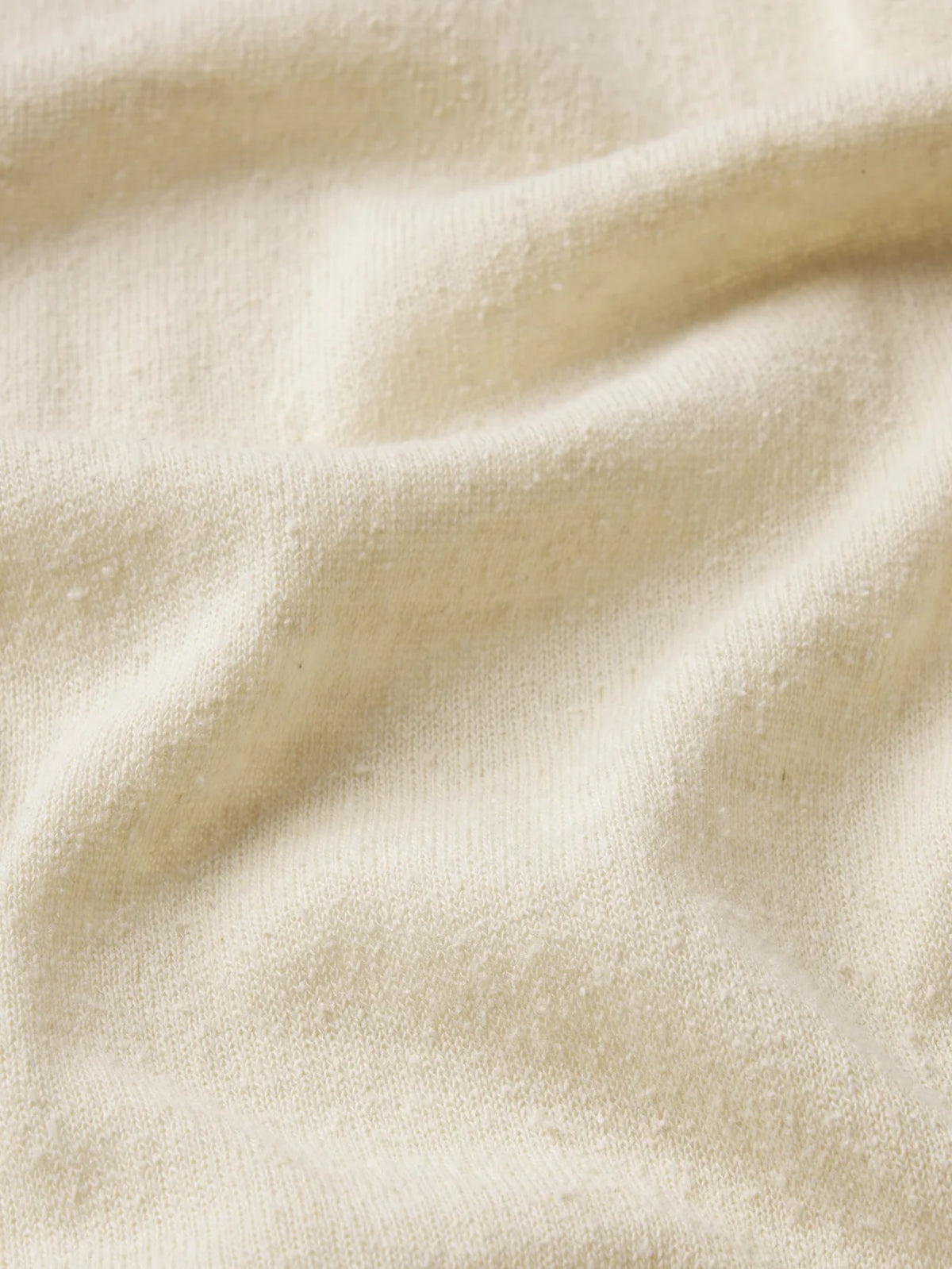 A close-up of The Knit Plus One Tee by FRAME shows its cream-colored, textured fabric with gentle folds and a nubby surface, capturing the relaxed luxury of this silk-inspired tee.