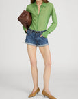 A person models FRAME’s The Collared Cardi in sheer green, styled with denim shorts, tan low-heeled shoes, and a large brown clutch against a plain light background.