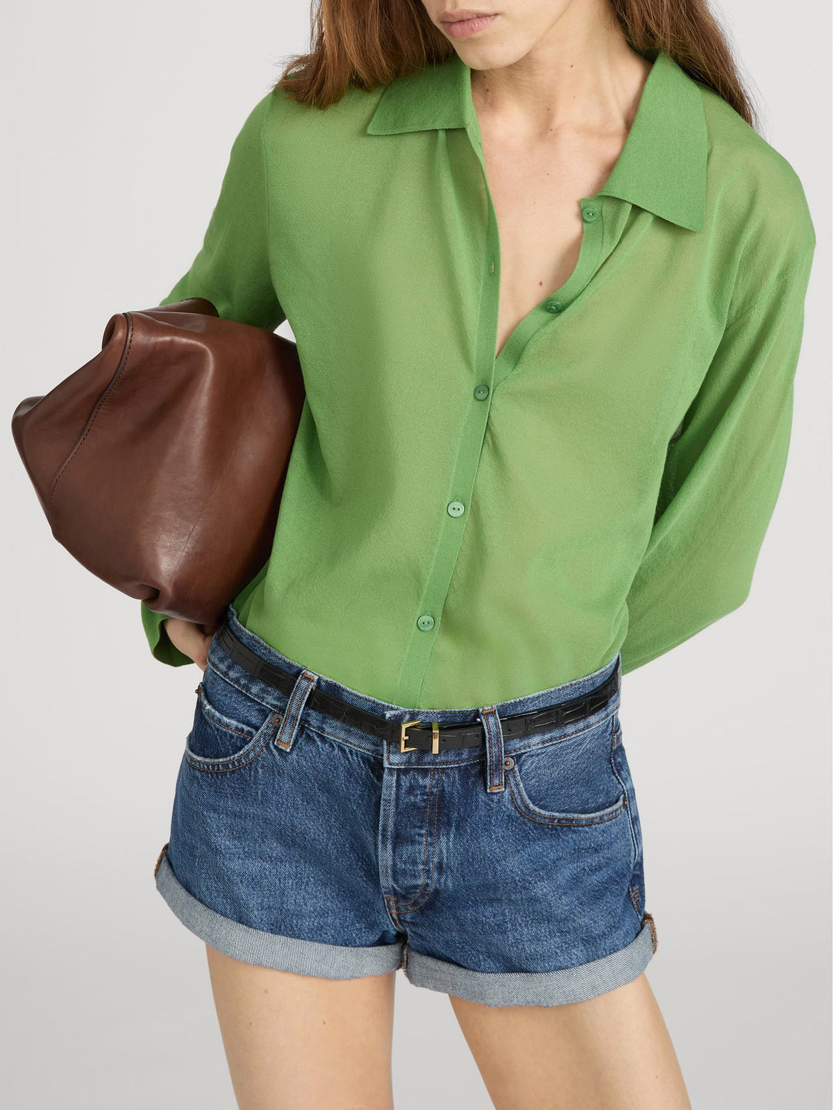 A woman wears The Collared Cardi by FRAME—a sheer green button-up styled like a lightweight knit—paired with denim shorts, a black belt, and carrying a brown leather clutch under her arm. Her head is slightly tilted, face partly visible.