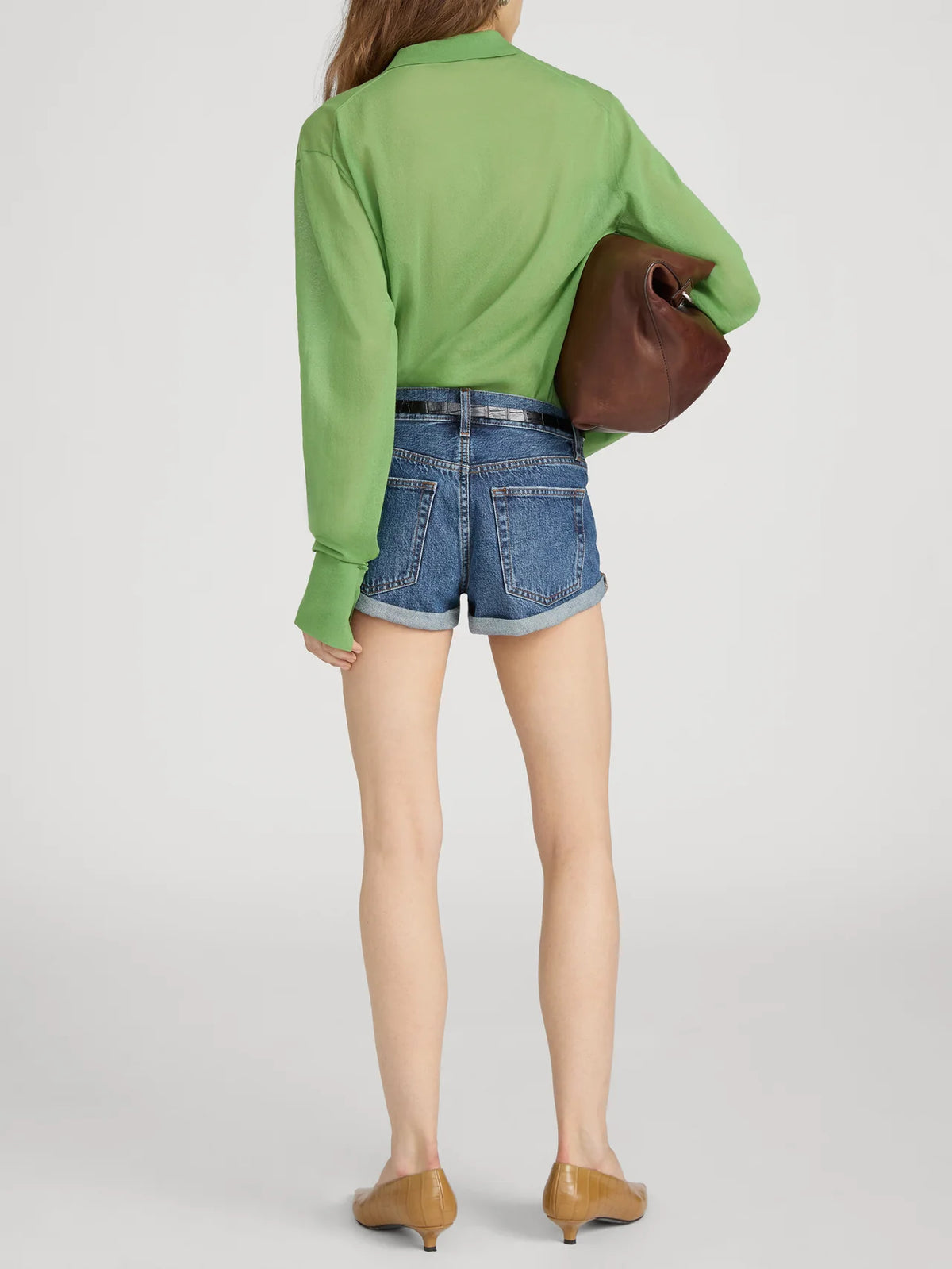 A person stands facing away, wearing FRAME’s The Collared Cardi in green with a slim fit, blue denim shorts, tan low-heeled shoes, and carrying a large brown clutch under one arm.