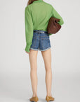 A person stands facing away, wearing FRAME’s The Collared Cardi in green with a slim fit, blue denim shorts, tan low-heeled shoes, and carrying a large brown clutch under one arm.