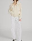 A person wearing FRAME's The Seersucker Track Jacket in light beige, paired with white wide-leg pants and black pointed-toe shoes, stands against a plain light gray background with their left hand in their pocket.