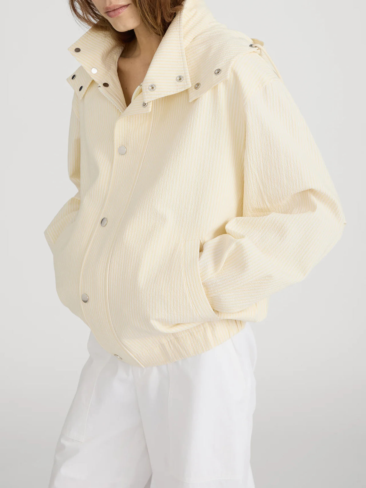 A person wears the FRAME Seersucker Track Jacket in light yellow, featuring snap buttons and a high collar, paired with white pants. Their hands are in their pockets, and their face is partially out of frame.