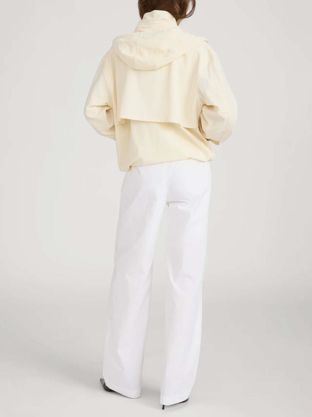 A person stands with their back to the camera, wearing FRAME's The Seersucker Track Jacket in light yellow with a vented back and loose white pants. The lightweight jacket pops against the plain white background.
