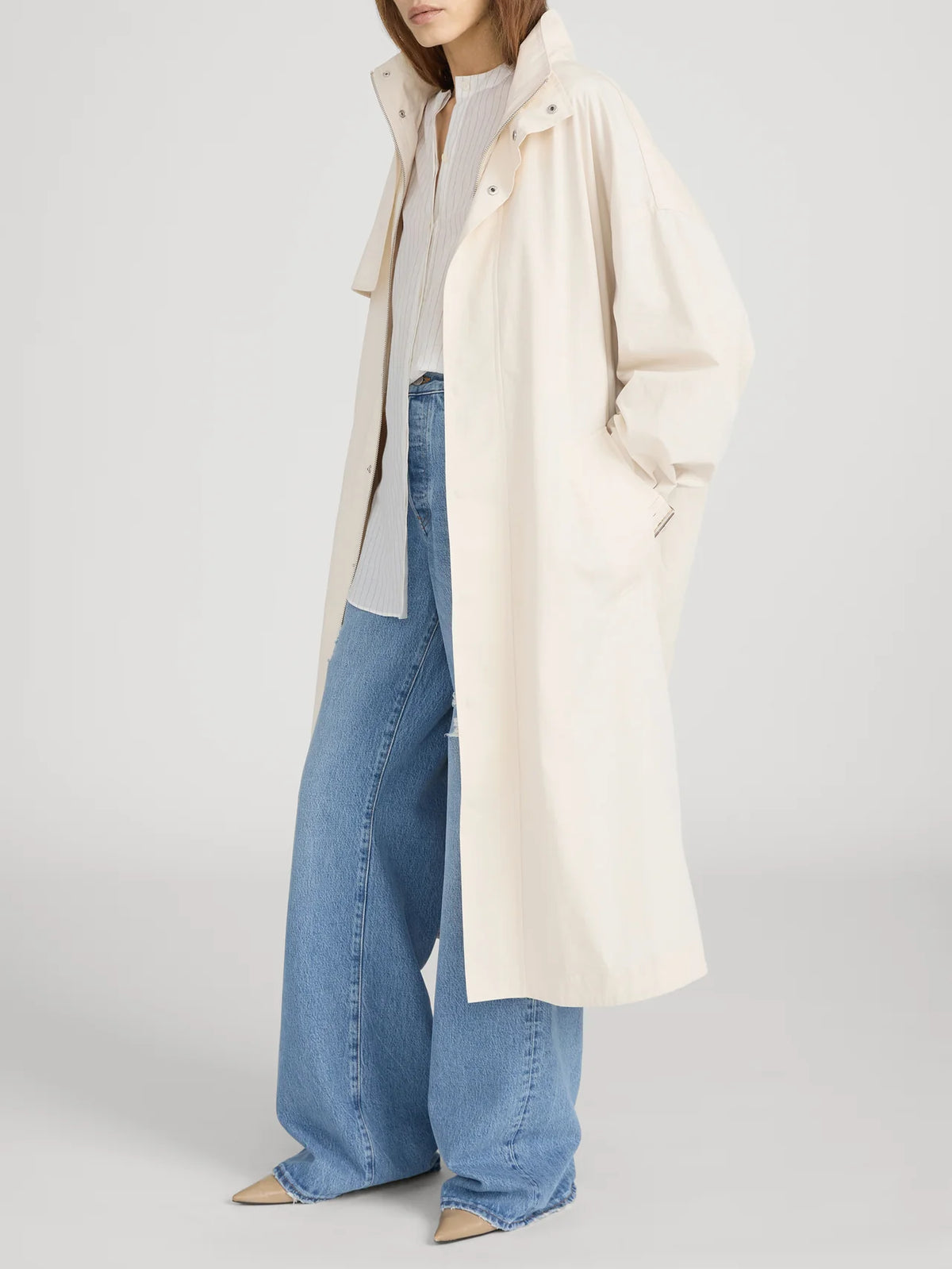 A woman wears The Field Trench by FRAME—a long, loose, cream cotton coat—over a light shirt and wide-leg blue jeans with beige pointed-toe shoes. One hand is in her pocket and her head is turned slightly to the side.