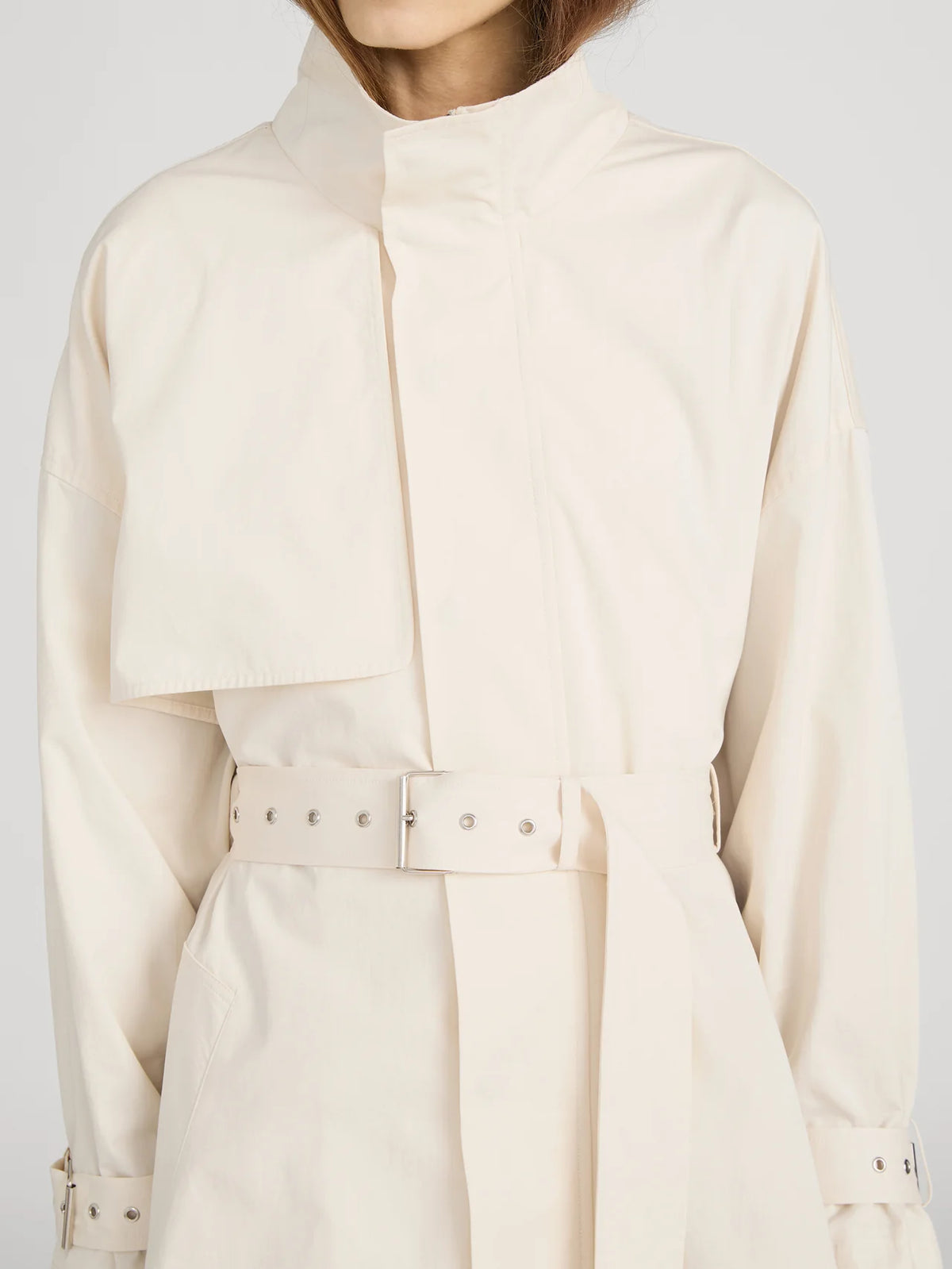 A person wears The Field Trench by FRAME, a cream cotton coat with a belted waist, high collar, chest flap detail, and metallic grommets. The face is partially out of frame for a chic, mysterious effect.