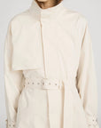 A person wears The Field Trench by FRAME, a cream cotton coat with a belted waist, high collar, chest flap detail, and metallic grommets. The face is partially out of frame for a chic, mysterious effect.