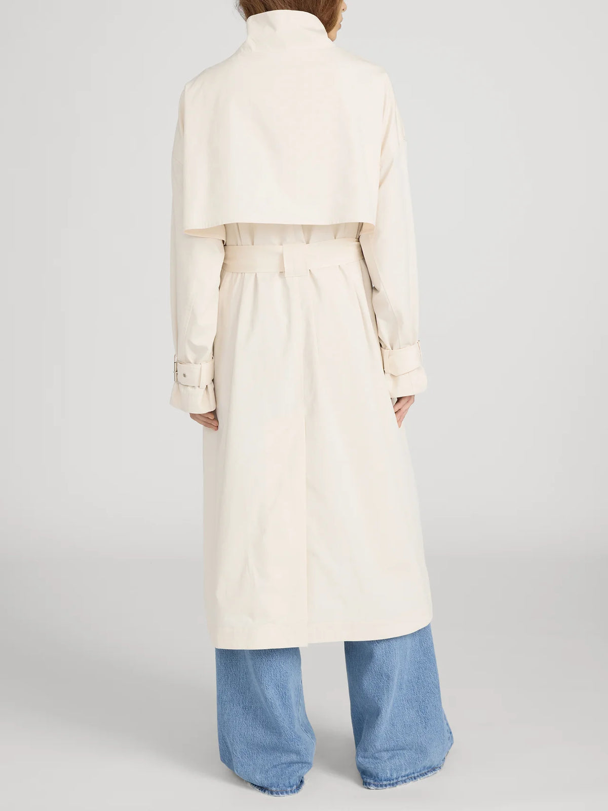 A person faces away from the camera wearing FRAME's The Field Trench in light beige with a belted waist and cape detail, paired with wide-leg blue jeans against a plain background.